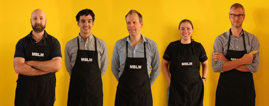 A group of men in black aprons standing in front of a yellow background.