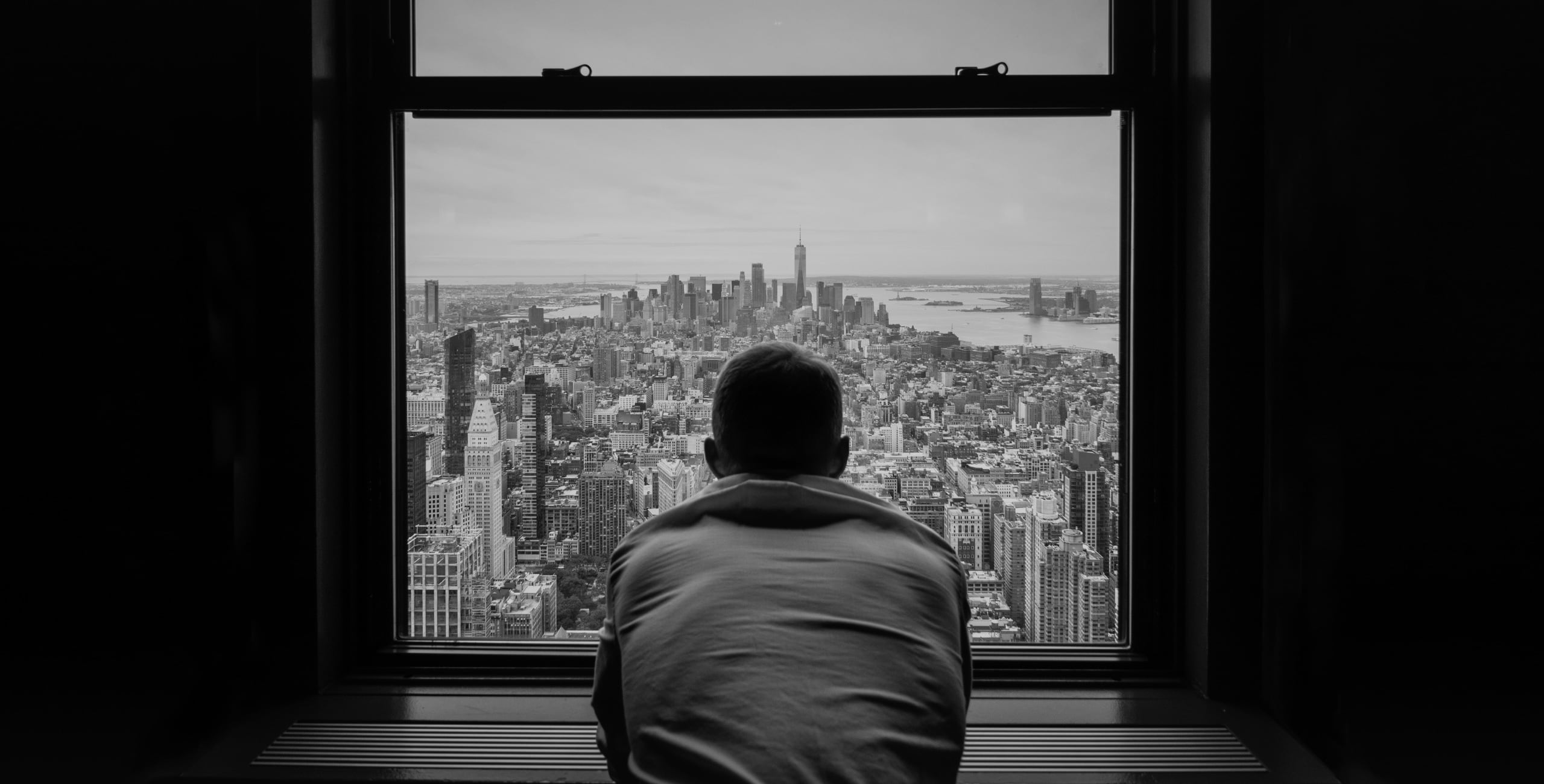 Black and white photo of a man looking out a window in new york city.