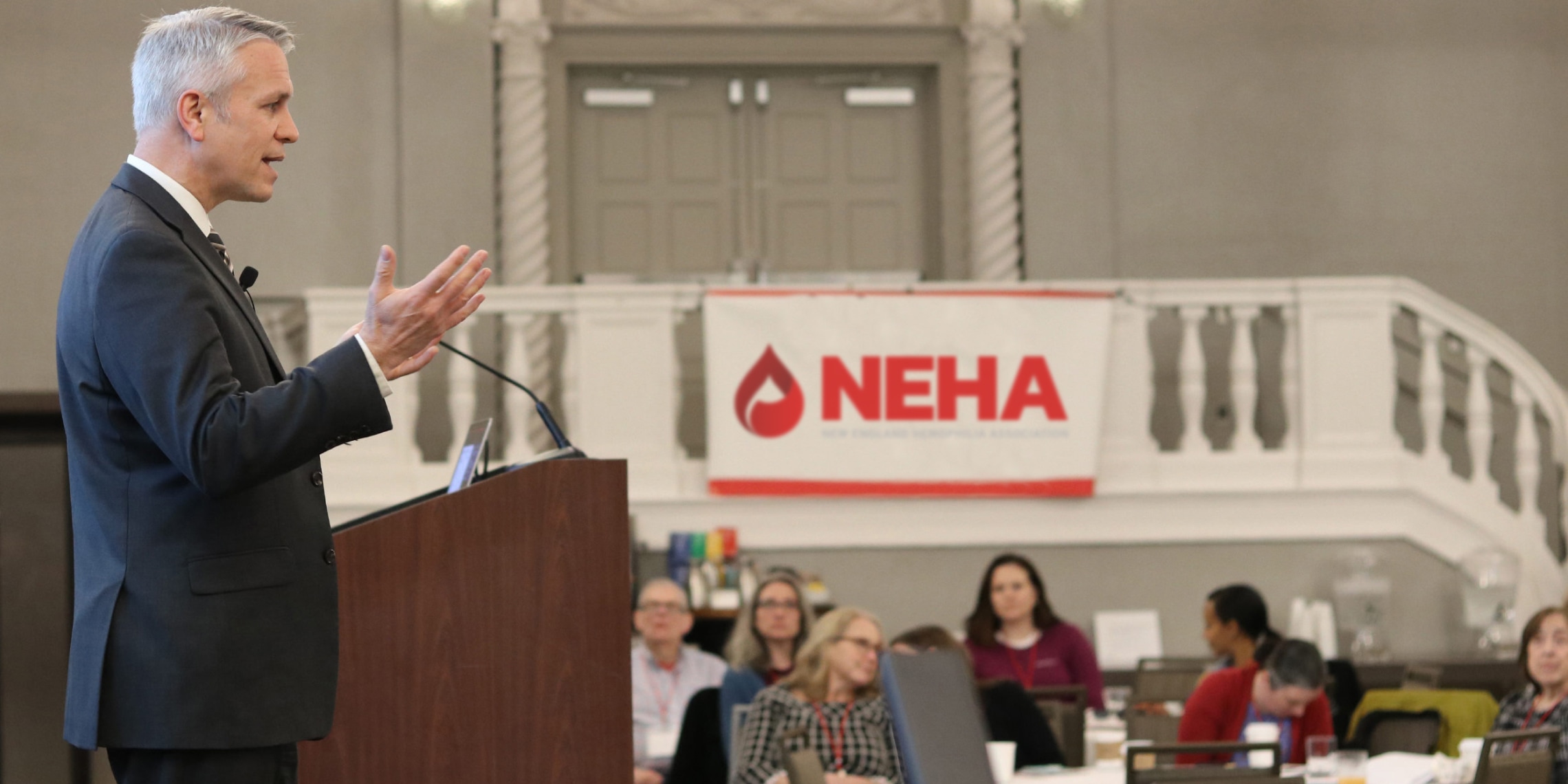 a person giving a speech in a conference room where a banner with Neha's logo can be seen.