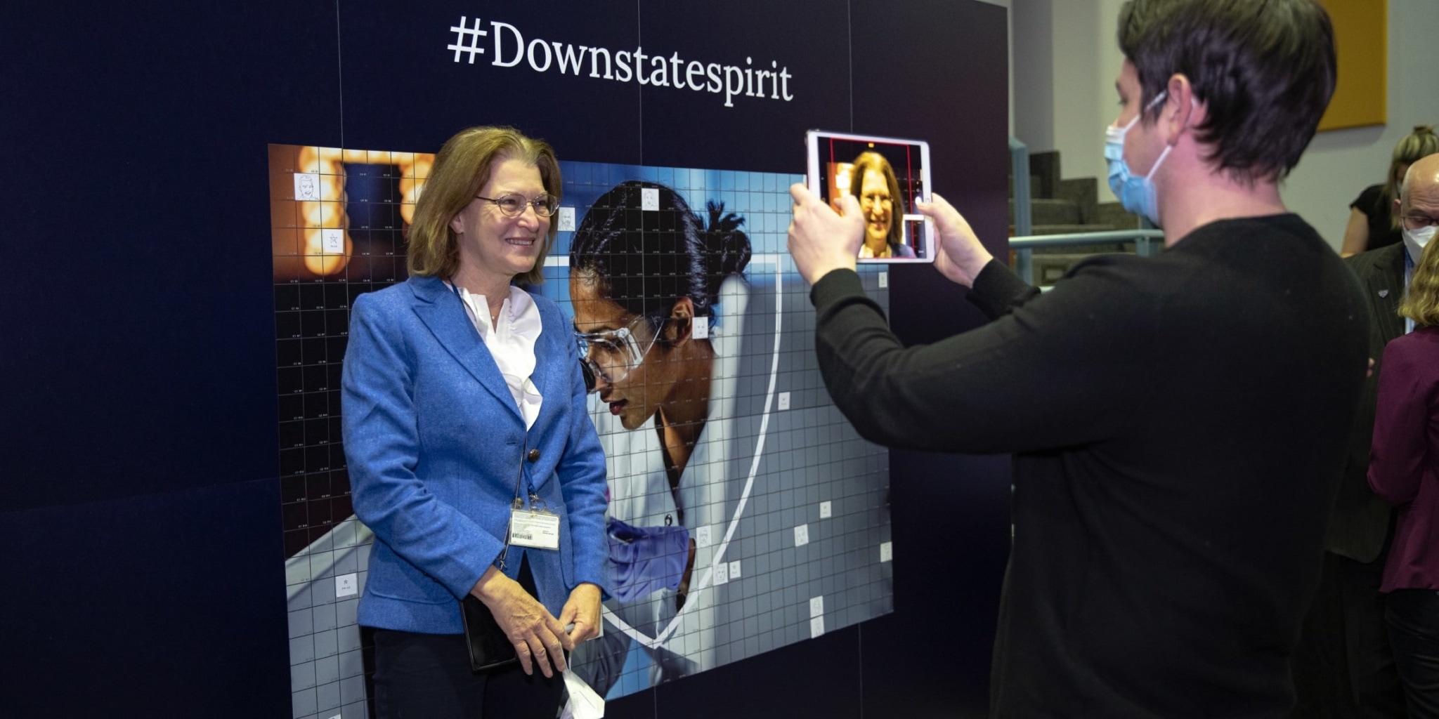 a college student taking a digital photo of a smiling woman standing in front of a wall with the new brand designed for SUNY.