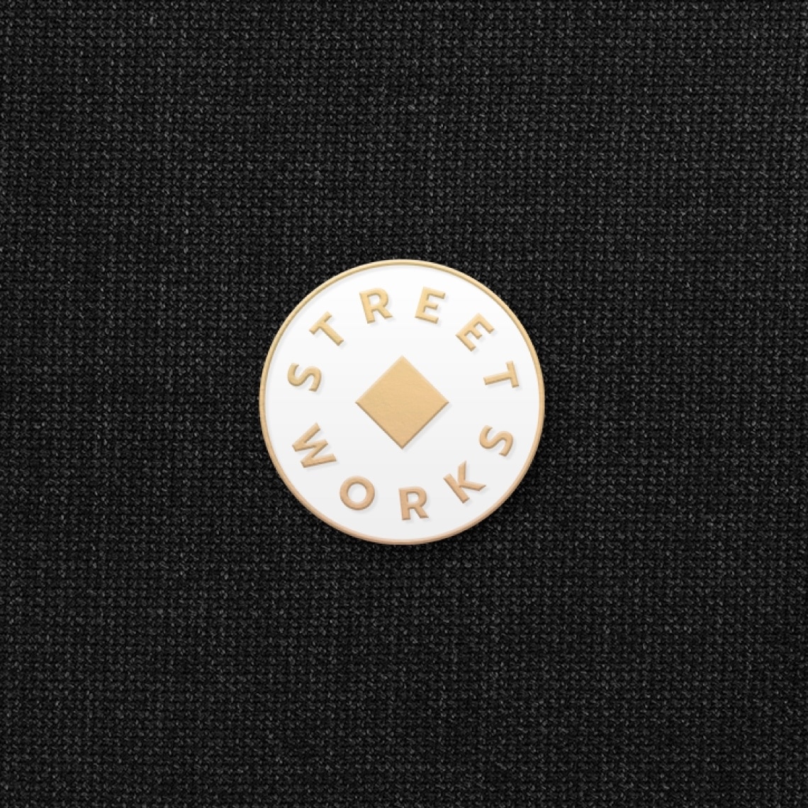 a white and gold pin with the logo of Street Works over a textured black fabric