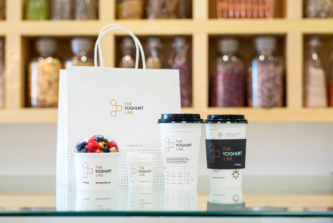 the design system developed for The Yoghurt Lab applied to branded materials such as shopping bags, ice cream containers, and beverage cups.