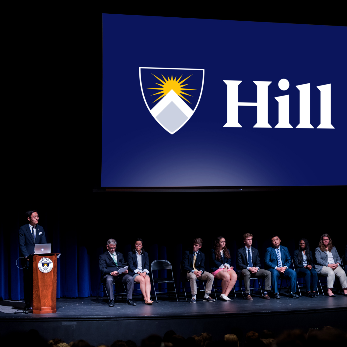 a group of people sitting on a stage with a large horizontal blue banner with the Hill School logo