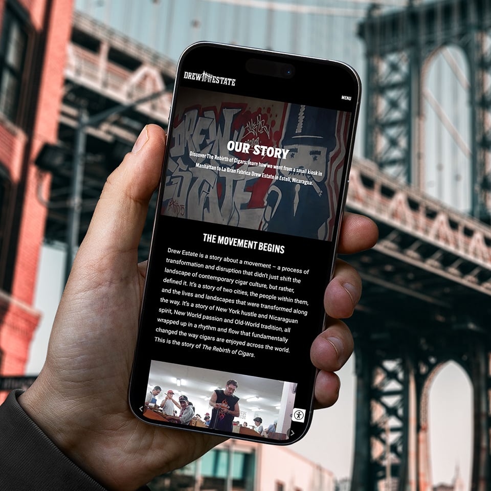 A hand holds a smartphone displaying the Drew Estate "Our Story" webpage, with a bridge structure blurred in the background.