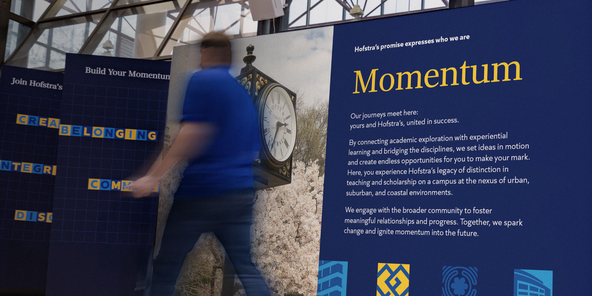 A person walking past large blue Hofstra University displays featuring the word "Momentum," descriptive text, and a photo of a campus clock.