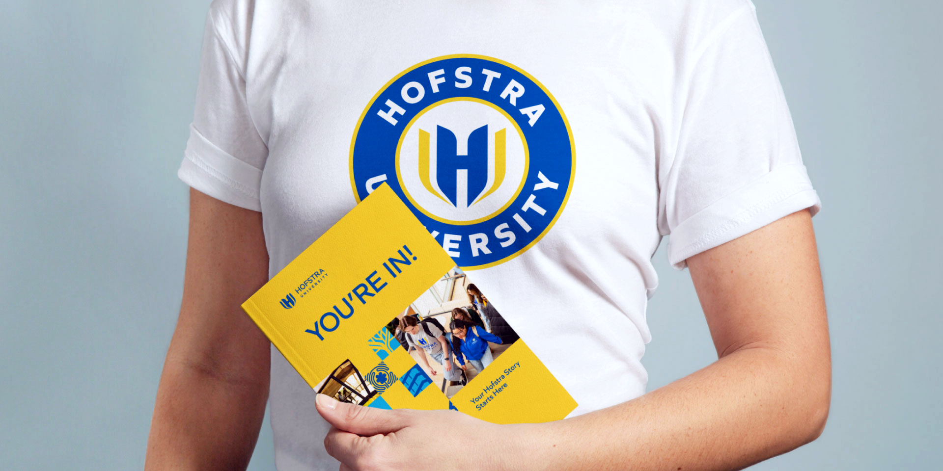 A person wearing a Hofstra University t-shirt holds a yellow welcome booklet that reads “YOU’RE IN!” with images of students and the university logo on the cover.