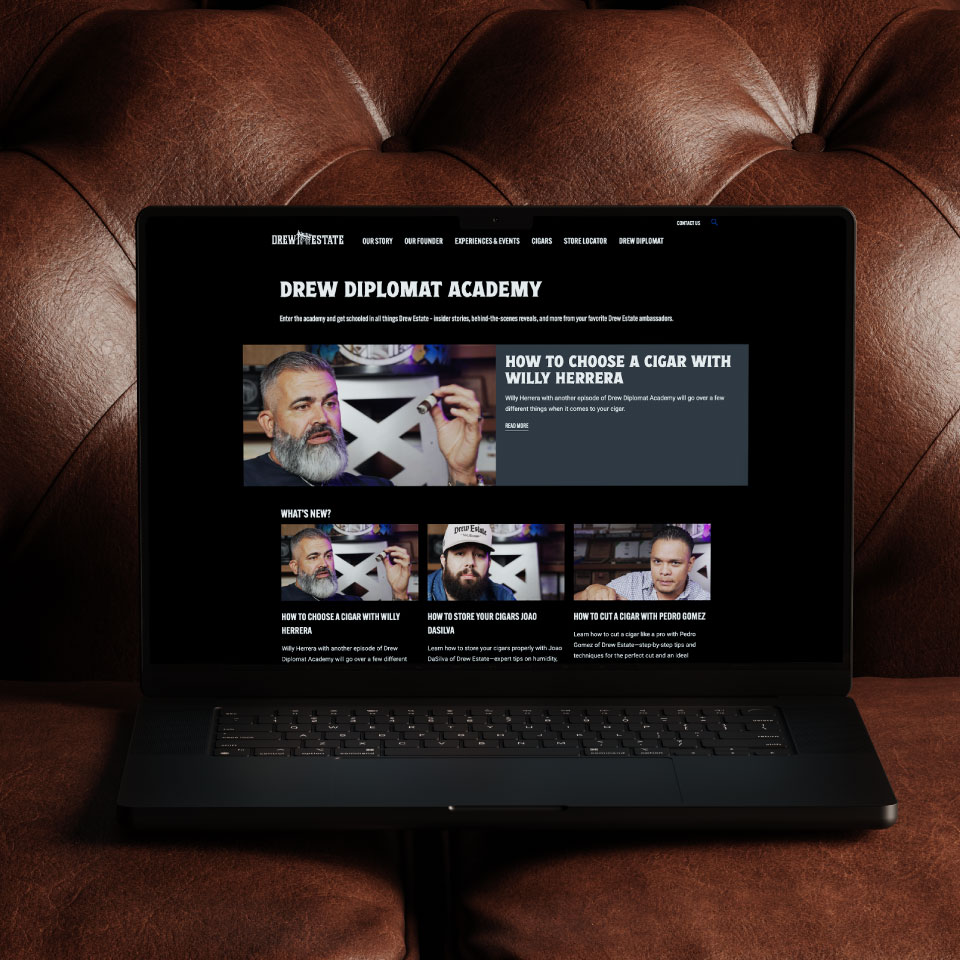 A laptop on a brown leather couch displays the Drew Diplomat Academy website, featuring videos on how to choose and enjoy cigars.