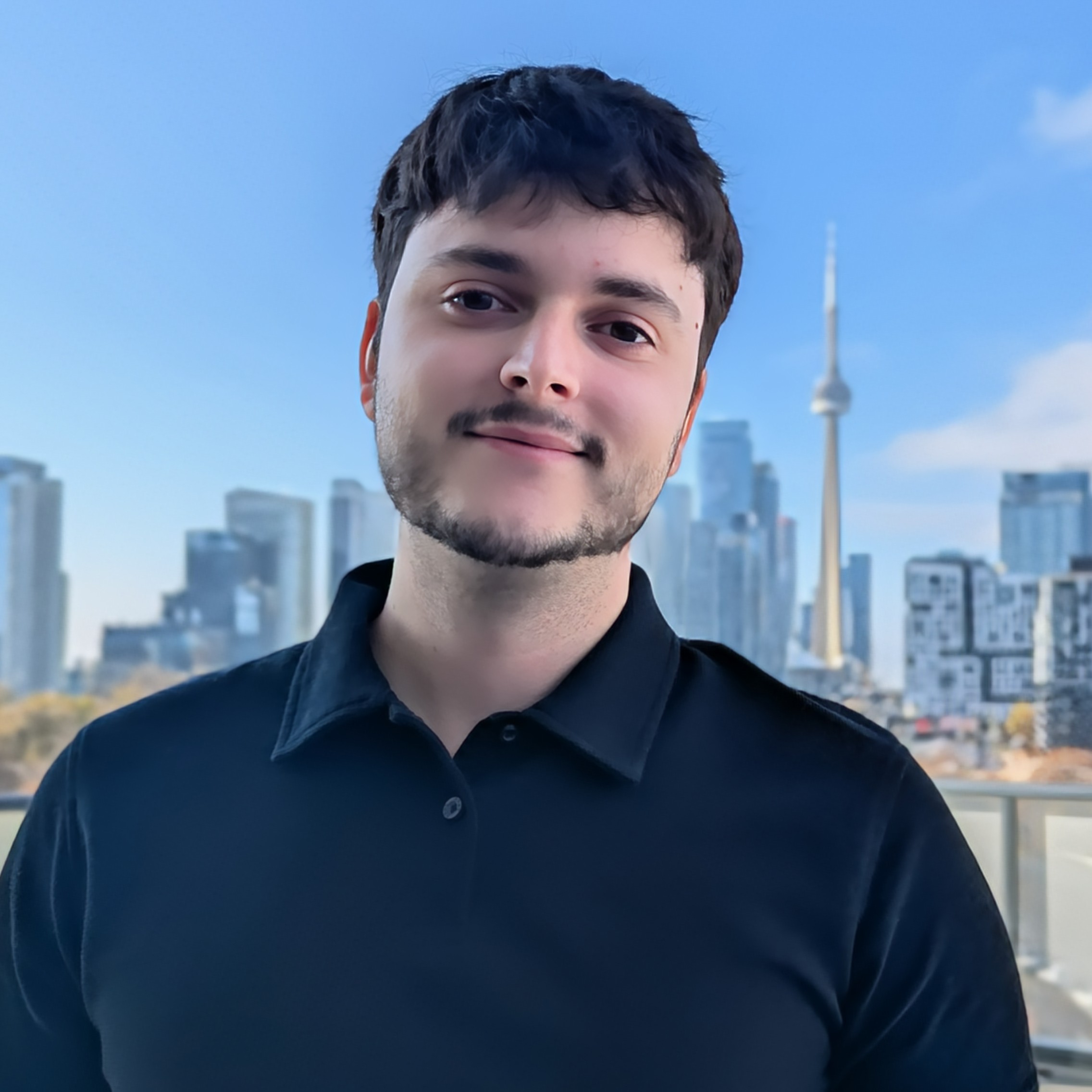 A young man wearing a black polo shirt stands on a balcony with a city skyline and the CN Tower in the background under a clear blue sky.
