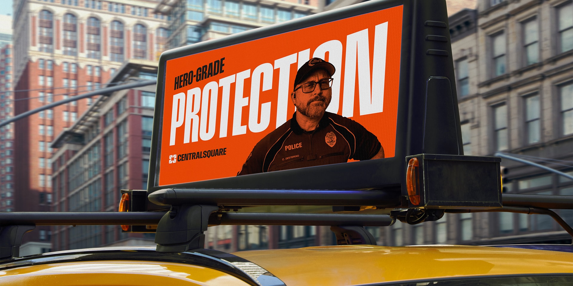 Digital billboard on top of a taxi displays an ad with a police officer and the text "HERO-GRADE PROTECTION" against an orange background in a city setting.