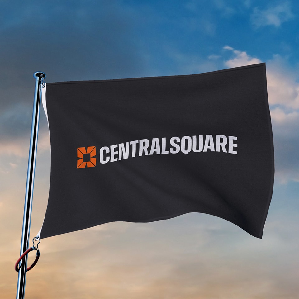 A black flag with the orange and white CentralSquare logo and text waving on a flagpole against a blue sky with clouds.