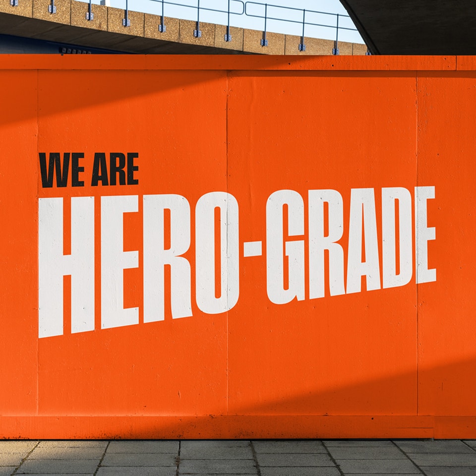 Bold white text on an orange wall reads "WE ARE HERO-GRADE," with the last two words in larger letters. A sidewalk and part of a bridge are visible above.
