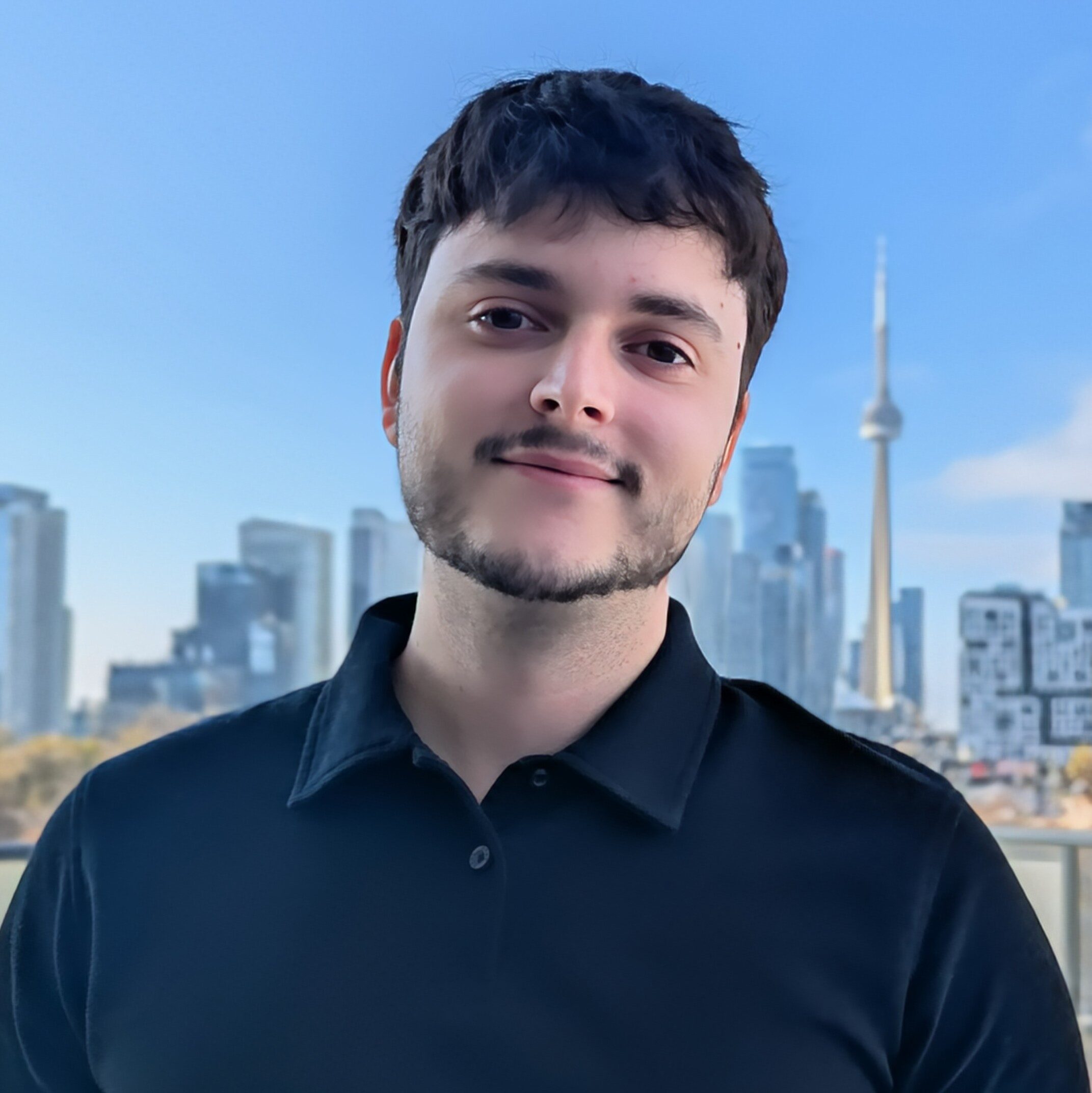 A person wearing a black collared shirt stands on a balcony with a city skyline and the CN Tower visible in the background on a clear day.