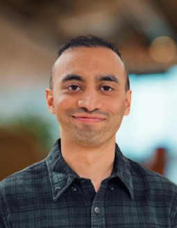 A person with short hair wearing a dark plaid shirt stands indoors with a blurred background.