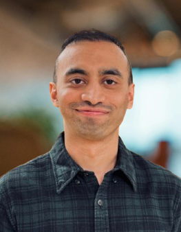 A person with short hair wearing a dark plaid shirt stands indoors with a blurred background.