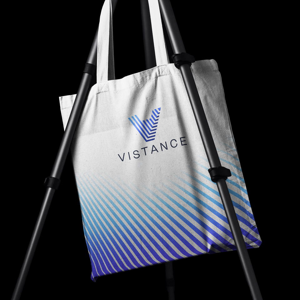 A white tote bag with blue diagonal lines and the word "VISTANCE" below a stylized "V" logo, hanging on a black tripod stand against a black background.