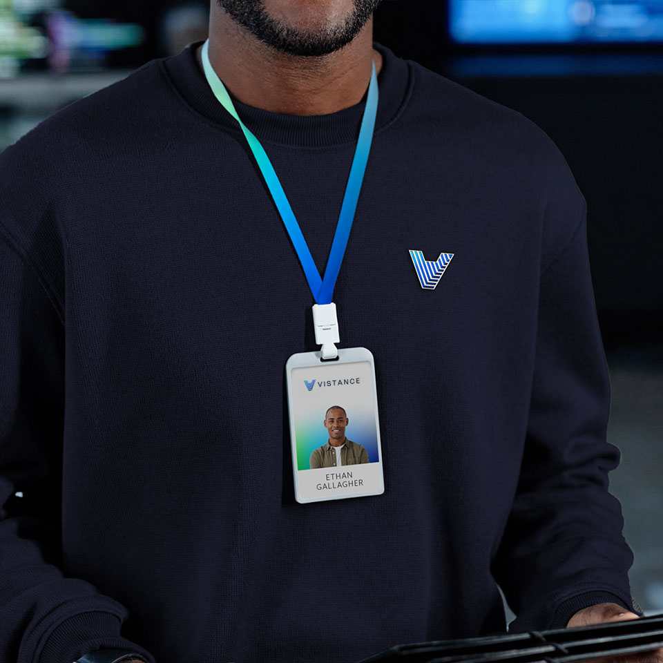 A person wearing a navy blue shirt with a "V" logo holds a tablet. An ID badge on a blue lanyard displays their photo and the name "Ethan Gallagher" with a "Vistance" logo.