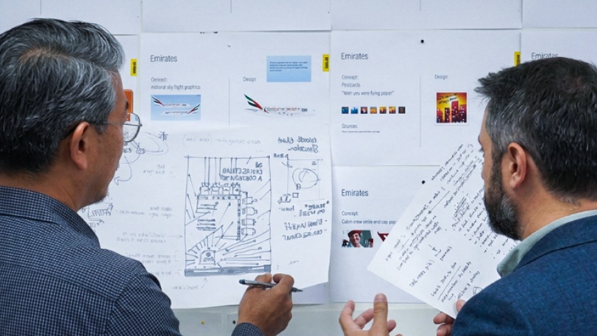 Two men discuss and review printed documents and sketches pinned to a whiteboard, which include diagrams, handwritten notes, and Emirates-branded project plans.