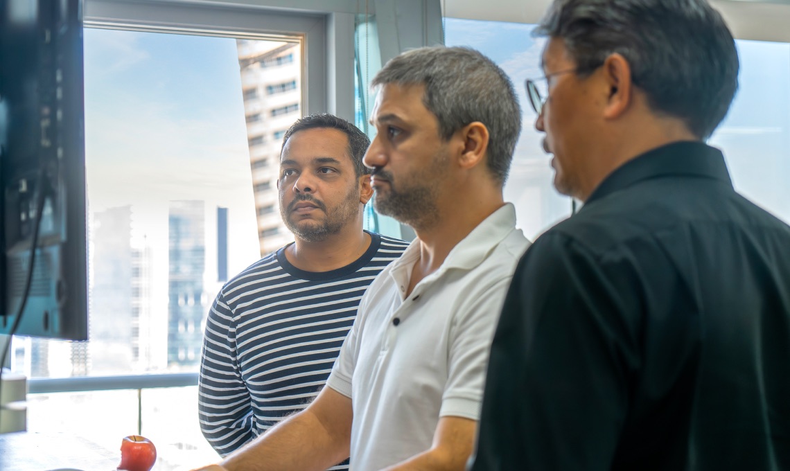 Three men stand together in an office, looking at a computer screen. One wears a striped shirt, another a white polo, and the third a black shirt. A window with city buildings is behind them.