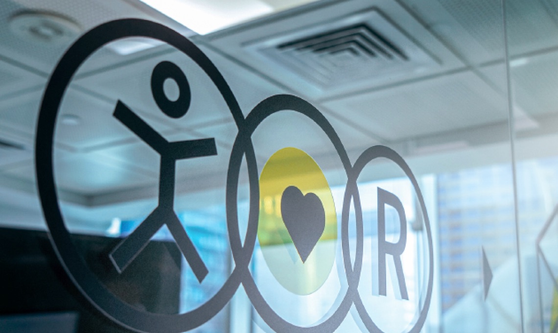 Frosted glass wall with symbols: a person, a heart, and a circled "R", in a modern office setting with ceiling panels and windows.