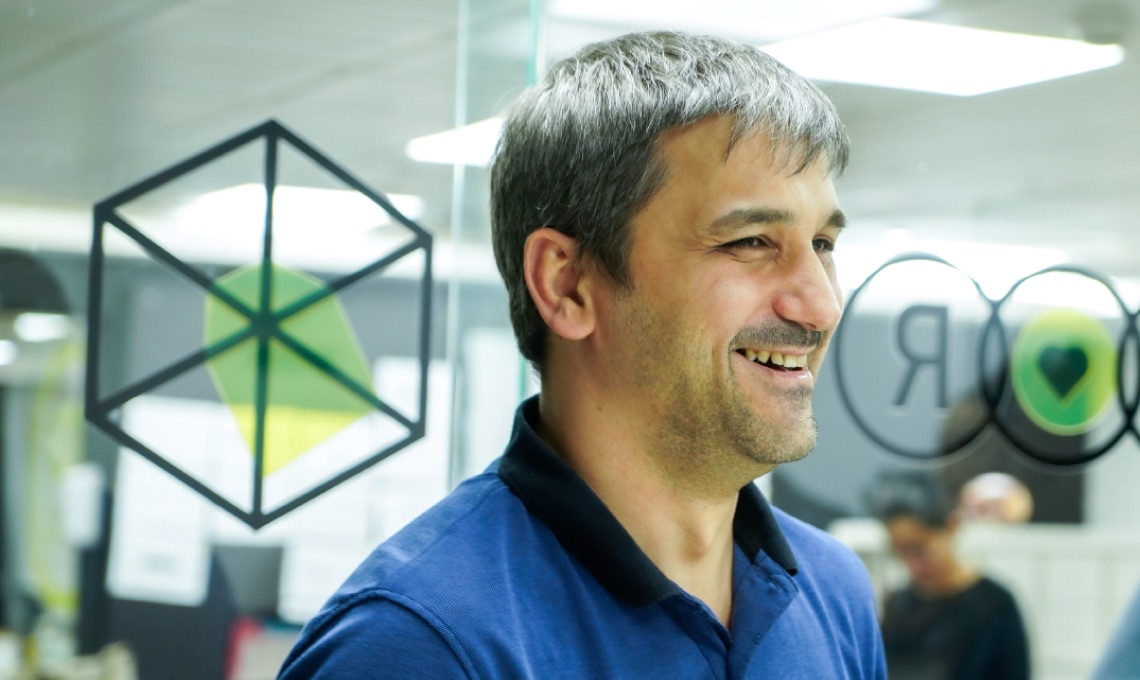 A man with short gray hair in a blue polo shirt smiles while standing indoors near glass panels with geometric designs.