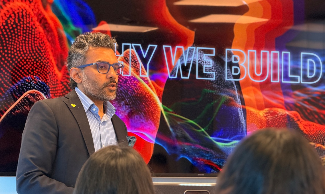 A man in a suit speaks in front of a colorful screen displaying the partial text "WHY WE BUILD" to an audience.