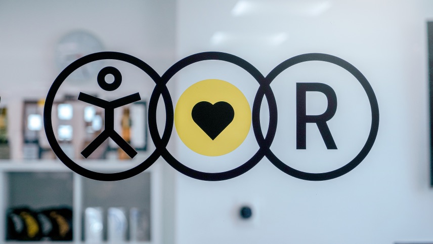 Three black-outlined circles on glass: the first has a stick figure, the second a yellow circle with a black heart, and the third contains the letter "R." Shelves are visible in the background.
