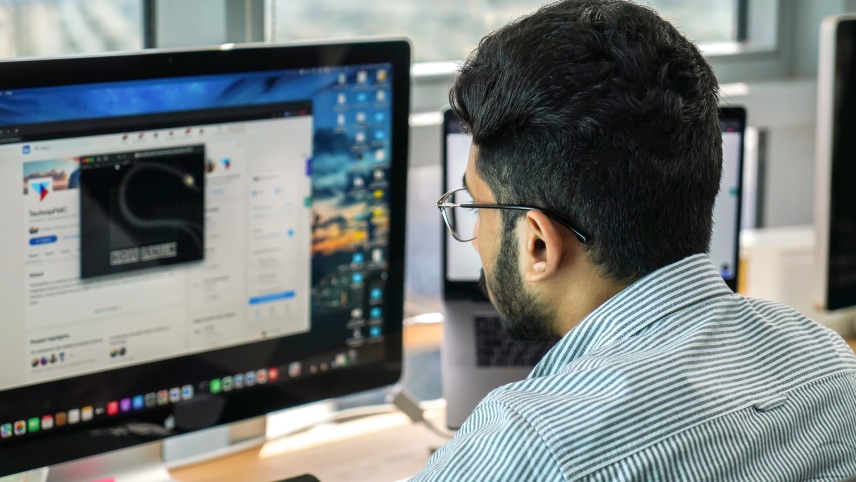 A man with glasses sits at a desk working on a desktop computer, displaying a webpage with various icons and text in an office setting.