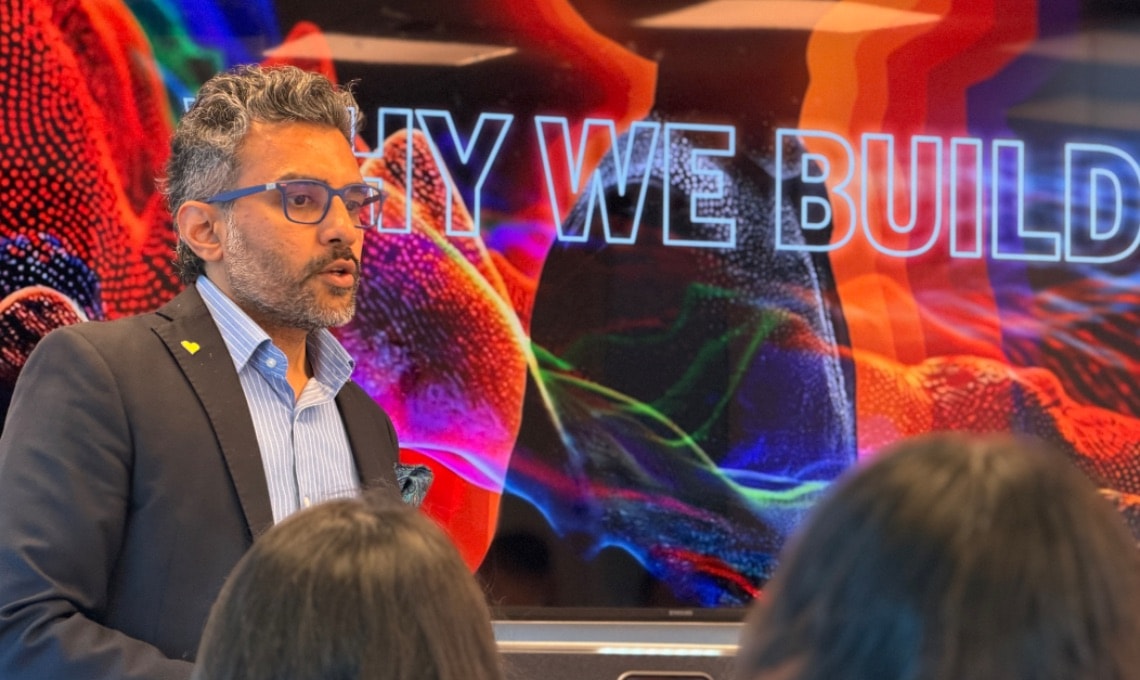A man in a suit speaks in front of a colorful screen displaying the words "WHY WE BUILD" to an audience.