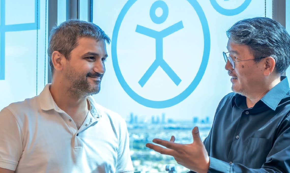Two men are having a conversation by a window with blue accessibility symbols on the glass. Cityscape is visible in the background.