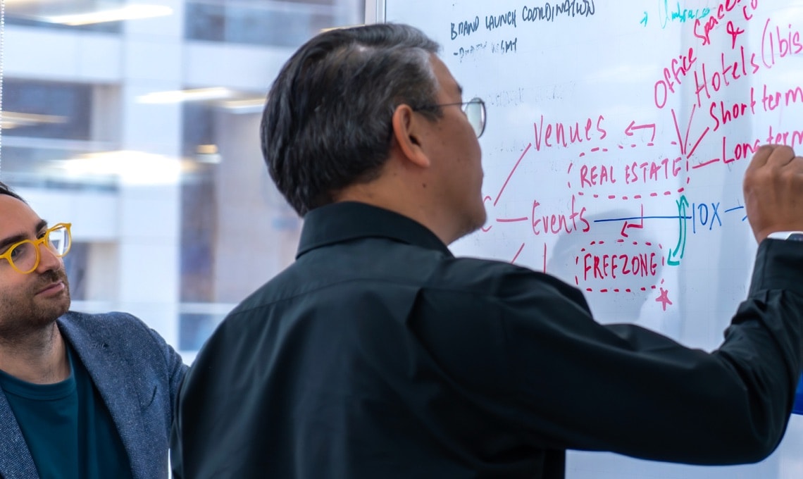 Two people collaborate at a whiteboard, one writing flowcharts and notes in red marker about venues, hotels, real estate, and events, while the other looks on attentively.