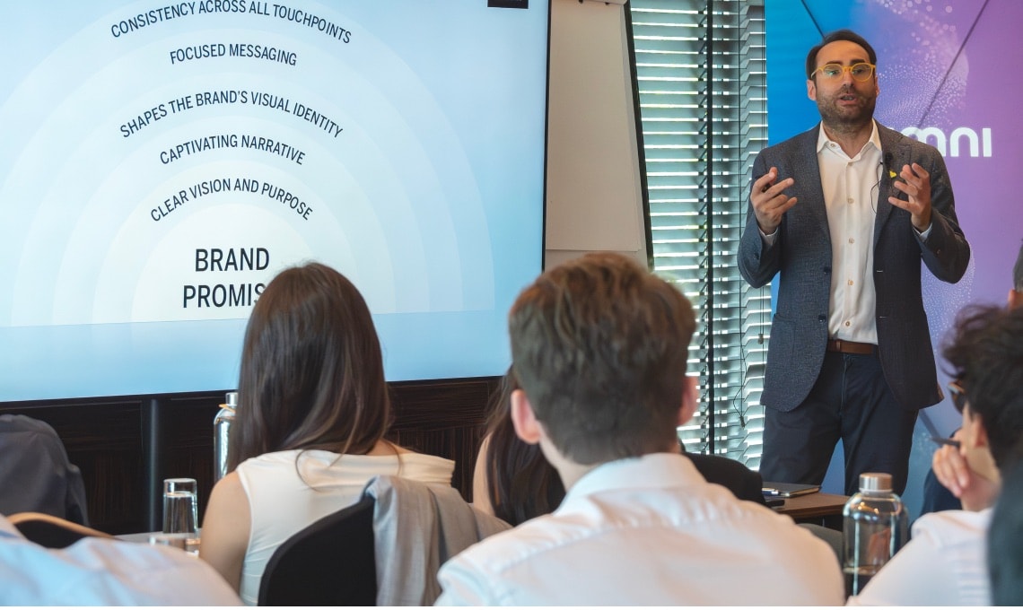 A man in a suit presents to an audience in a conference room. A slide on the screen lists steps that lead to a brand promise.