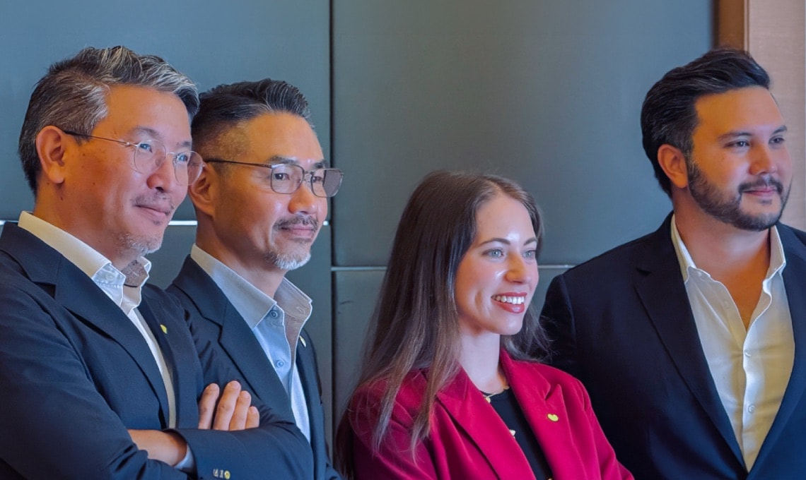 Four people wearing business attire stand together indoors, smiling and looking slightly to the right of the camera.