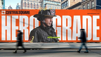 Large billboard on a city street shows a firefighter in uniform with the words “Hero Grade” behind him; two blurred pedestrians walk past.